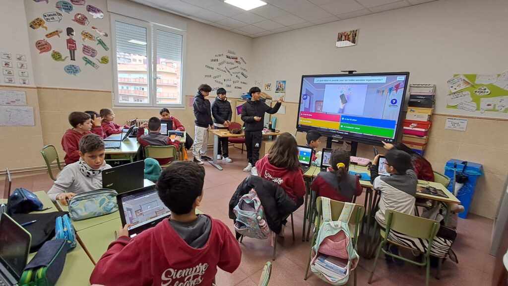 Alumnado de Educación Primaria participa en una sesión sobre uso seguro de las tecnologías mientras estudiantes mayores actúan como ciberayudantes utilizando una pantalla digital en el aula