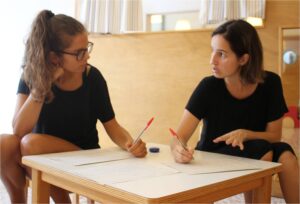 Dos maestras sentadas a la misma mesa conversa sobre el trabajo conjunto que han realizado en el aula