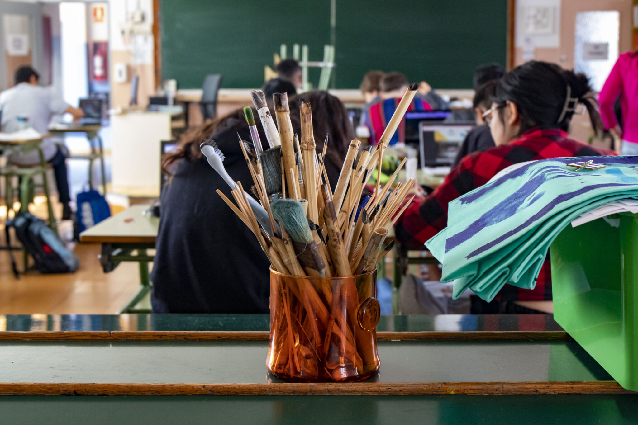 bote de lápices en primer plano. Tras él, varios estudiantes sentados en sus mesas en un aula de instituto