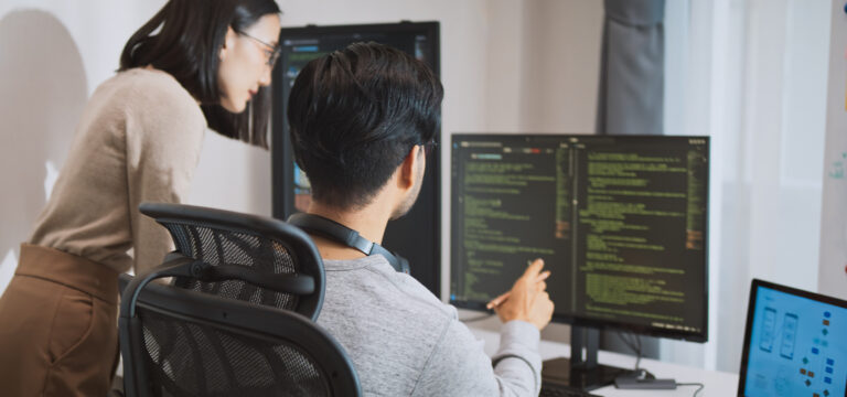 Dos personas charlan sobre el código de programación que aparece en sendas pantallas frente ellas.