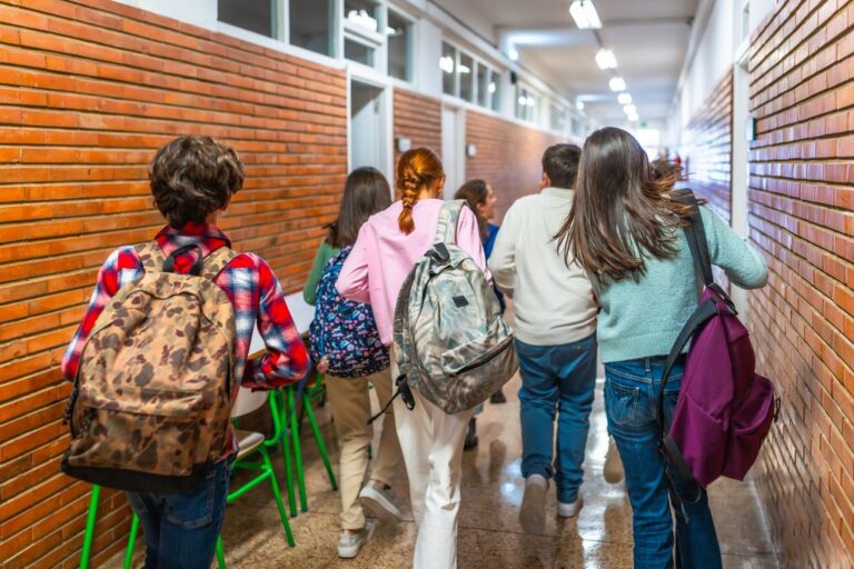 Un grupo de niñas y niños caminan con sus mochilas por el pasillo de un colegio. Dan la espalda a la cámara