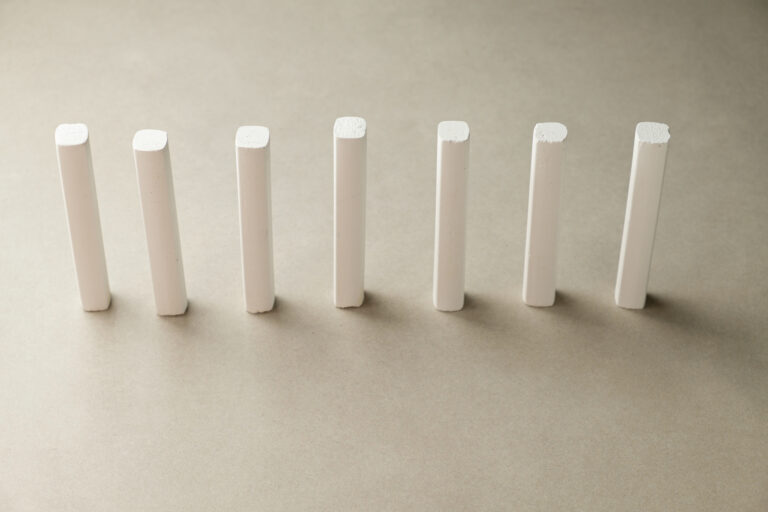 Seven white chalk sticks stand upright in a neat line on a subtle gray background, showcasing their smooth surfaces and clean edges in perfect symmetry under soft lighting.