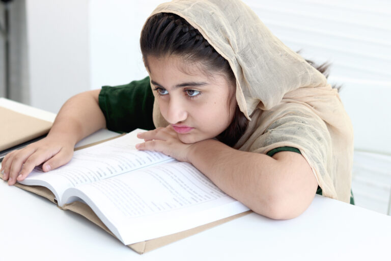 Adorable Pakistani Muslim sleepy girl with beautiful eyes wearing hijab, feeling bored and tired during studying and doing homework on table, student kid reading book on white background.